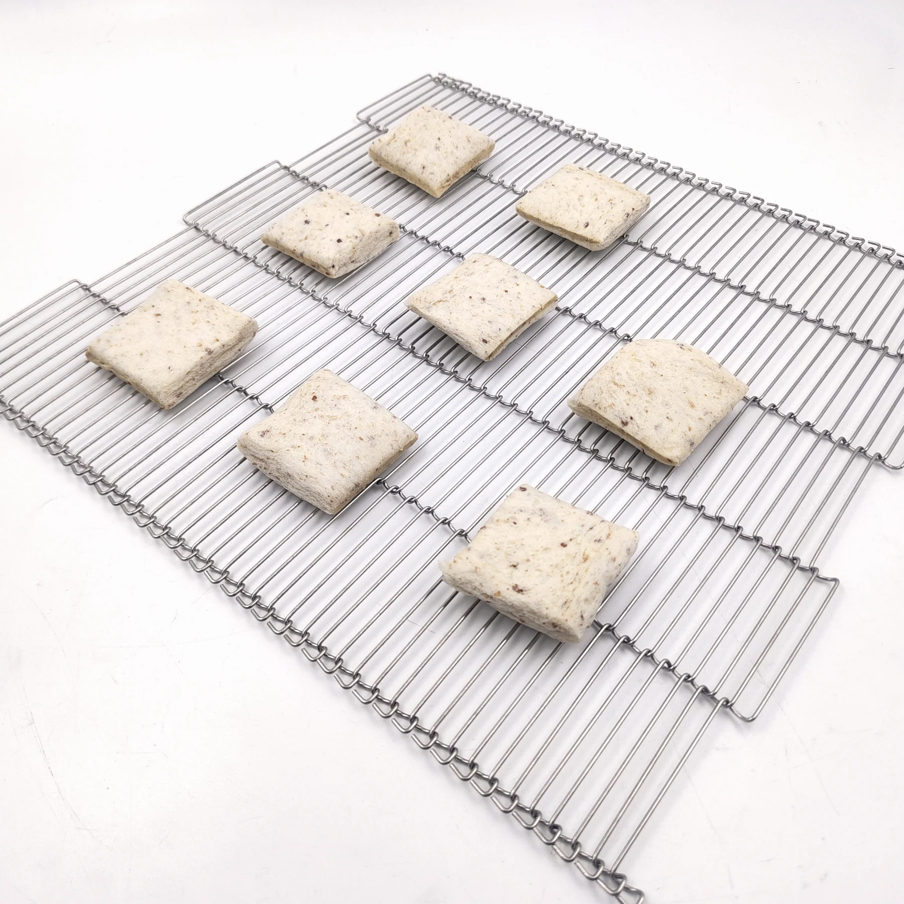 Square prepared food portions arranged across an open ladder-style metal conveyor belt.